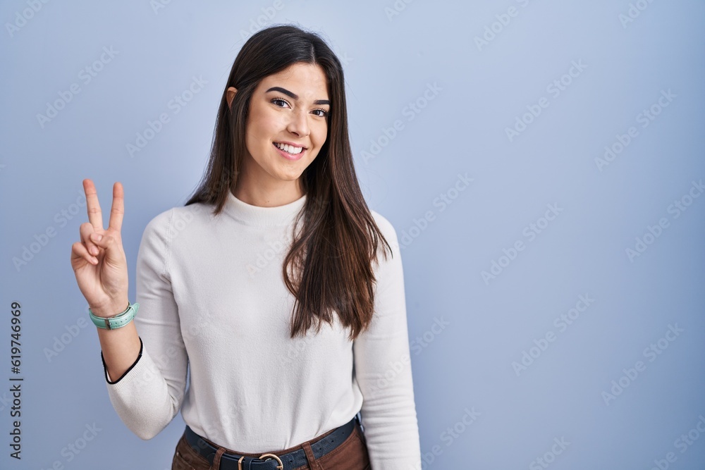 Young brunette woman standing over blue background showing and pointing up with fingers number two while smiling confident and happy. Young brunette woman standing over blue background showing and pointing up with fingers number two while smiling confident and happy.