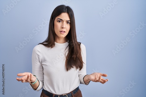 Young brunette woman standing over blue background clueless and confused with open arms, no idea concept.