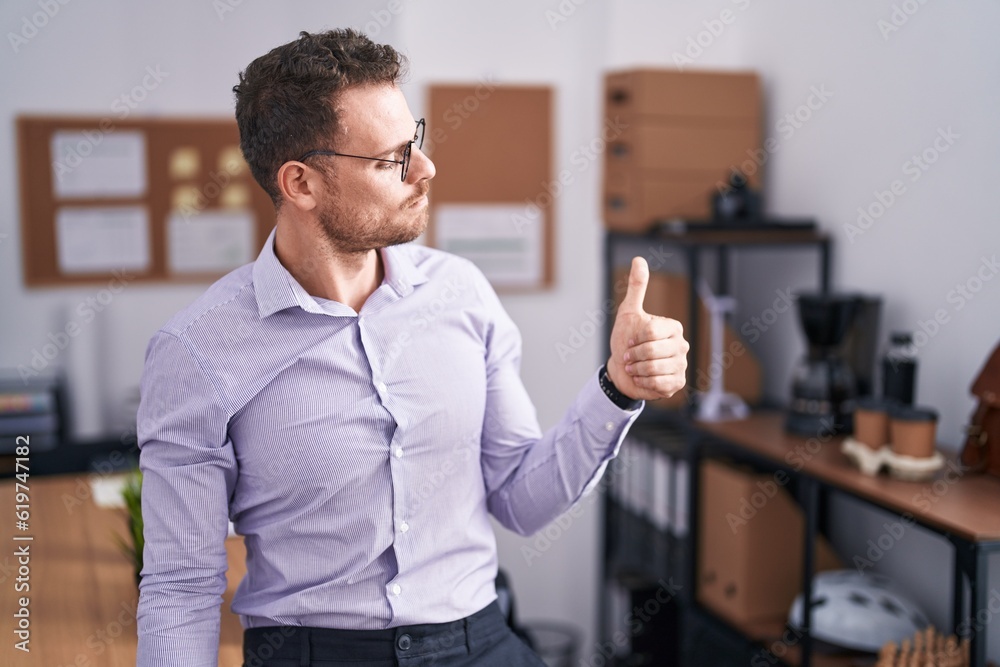 Young hispanic man at the office looking proud, smiling doing thumbs up gesture to the side Young hispanic man at the office looking proud, smiling doing thumbs up gesture to the side
