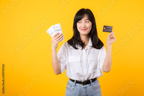 happy successful confident young asian woman happy smile wearing white shirt and denim jean holding cash money and credit card standing over yellow background. millionaire business, shopping concept.