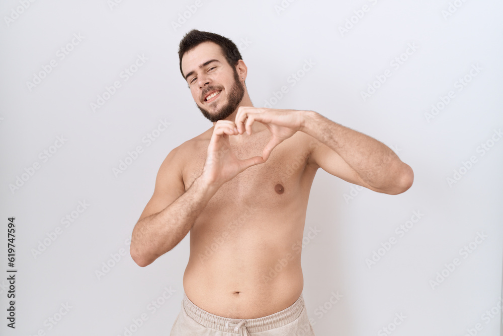 Young hispanic man standing shirtless over white background smiling in love doing heart symbol shape with hands. romantic concept.