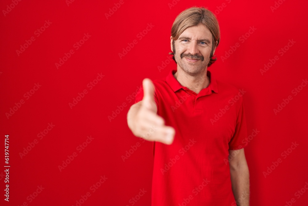 Caucasian man with mustache standing over red background smiling friendly offering handshake as greeting and welcoming. successful business. Caucasian man with mustache standing over red background smiling friendly offering handshake as greeting and welcoming. successful business.