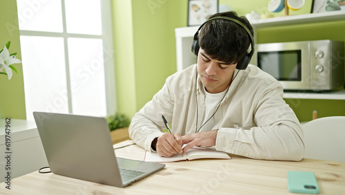 Young hispanic man student using computer taking notes wearing headphones at dinning room
