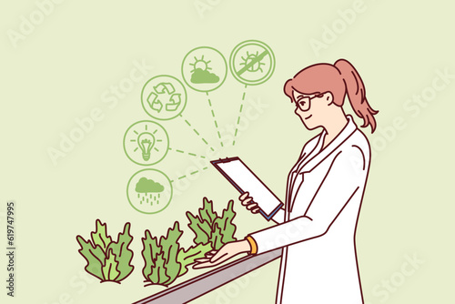 Woman works in hydroponic plant farm growing plants in biological research laboratory. Happy girl in white coat is studying herbs with beneficial properties working in scientific farm.