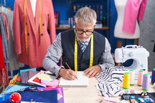 Middle age grey-haired man tailor drawing on notebook at clothing factory