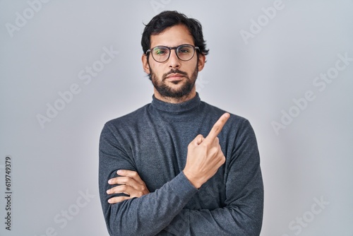 Handsome latin man standing over isolated background pointing with hand finger to the side showing advertisement, serious and calm face © Krakenimages.com