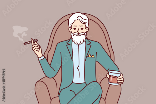 Elderly man aristocrat sits in armchair in expensive formal suit and drinks brandy with cigar. Millionaire aristocrat with gray mustache and beard poses haughtily with glass of alcohol photo