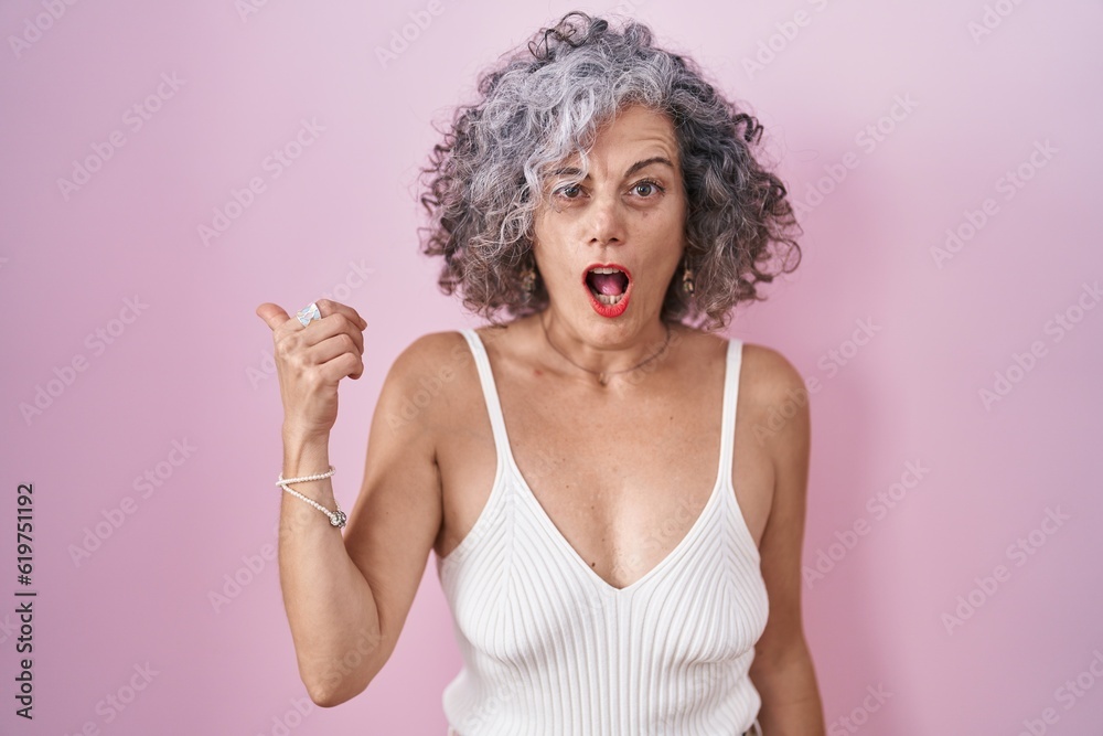 Middle age woman with grey hair standing over pink background surprised pointing with hand finger to the side, open mouth amazed expression.