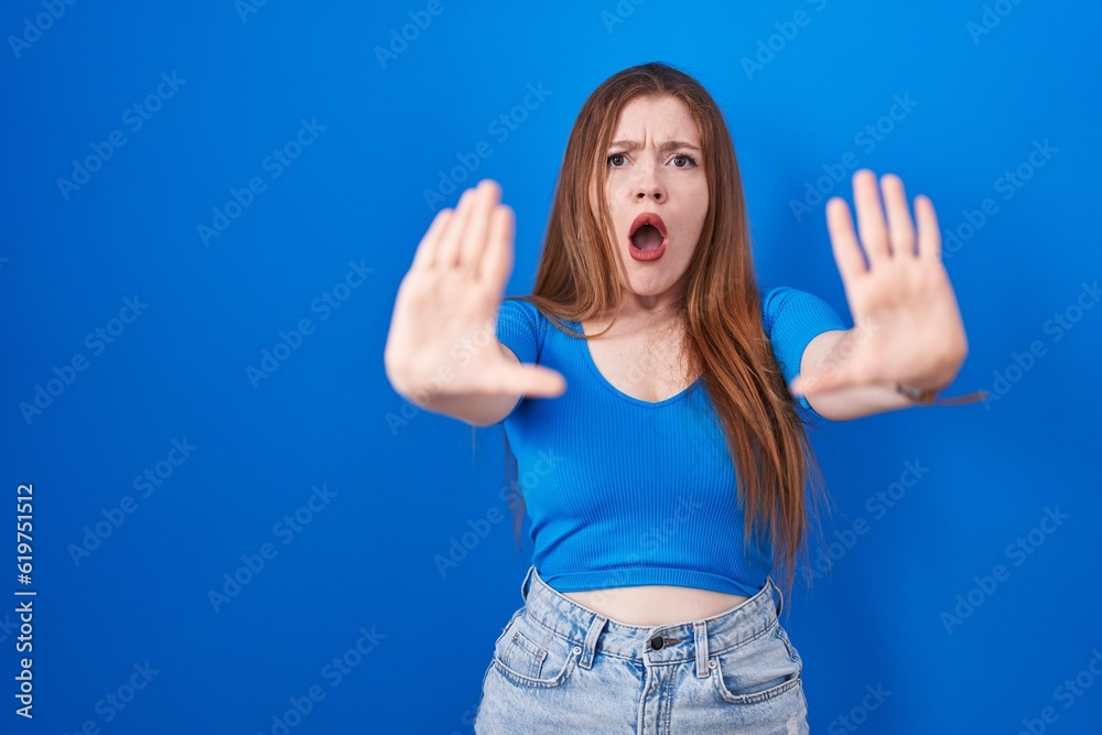 Redhead woman standing over blue background doing stop gesture with hands palms, angry and frustration expression Redhead woman standing over blue background doing stop gesture with hands palms, angry and frustration expression
