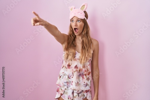Young caucasian woman wearing sleep mask and pajama pointing with finger surprised ahead, open mouth amazed expression, something on the front