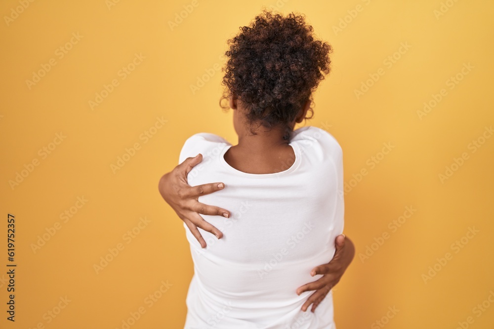 Young hispanic woman with curly hair standing over yellow background hugging oneself happy and positive from backwards. self love and self care Young hispanic woman with curly hair standing over yellow background hugging oneself happy and positive from backwards. self love and self care
