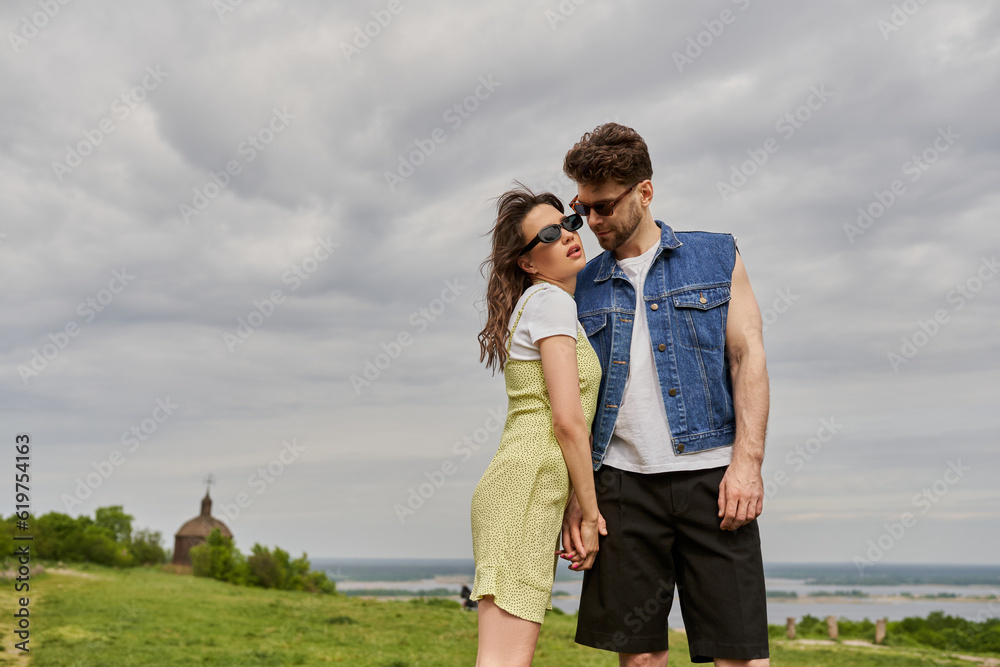 Stylish bearded man in denim vest touching hand of romantic girlfriend in sunglasses and sundress while standing together with rural setting at background, countryside retreat concept Stylish bearded man in denim vest touching hand of romantic girlfriend in sunglasses and sundress while standing together with rural setting at background, countryside retreat concept