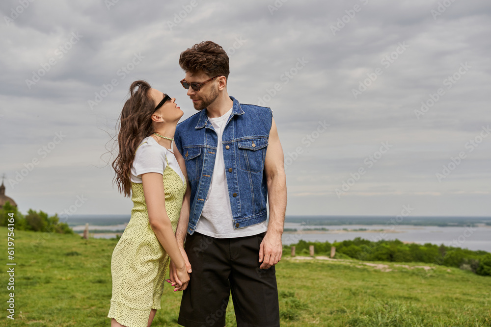 Romantic brunette woman in sunglasses and sundress holding hand of bearded boyfriend in denim vest and standing together with rural landscape and cloudy sky at background, countryside retreat concept Romantic brunette woman in sunglasses and sundress holding hand of bearded boyfriend in denim vest and standing together with rural landscape and cloudy sky at background, countryside retreat concept