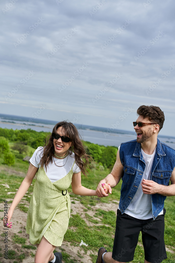 Positive brunette man in sunglasses and denim vest holding hand of cheerful girlfriend in stylish sundress and spending time with nature at background, countryside leisurely stroll, tranquility