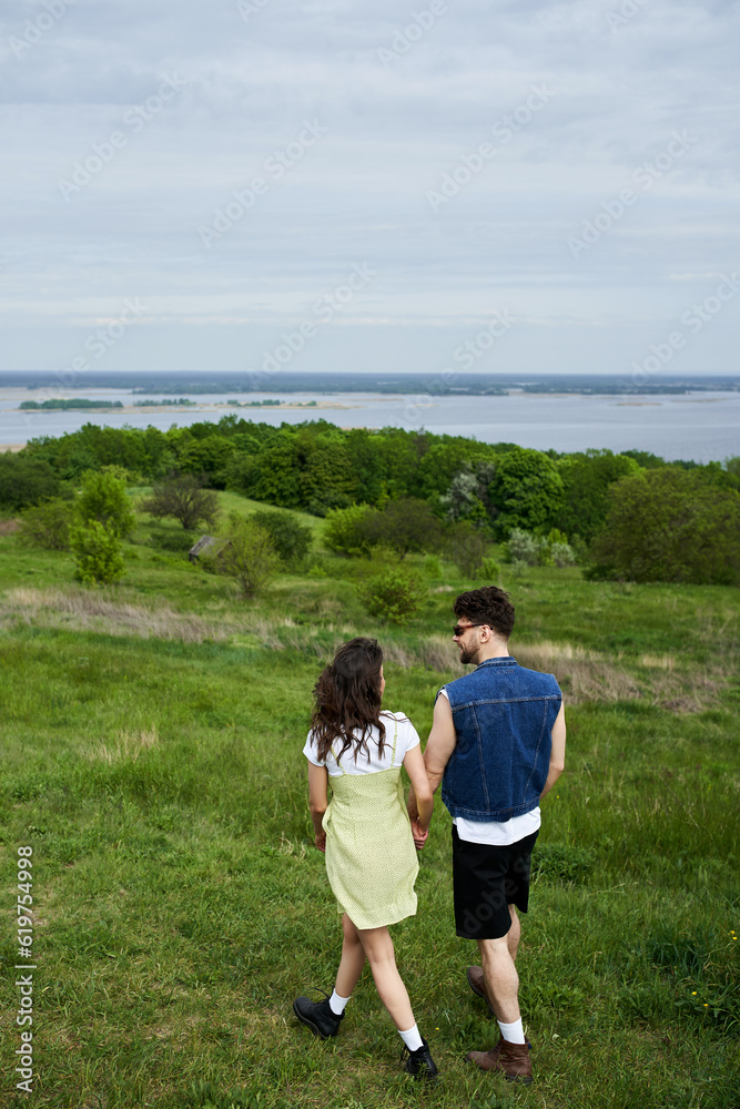 Brunette man in sunglasses and denim vest holding hand of stylish girlfriend in boots and sundress and walking together on field with green grass at summer, countryside adventure and love story Brunette man in sunglasses and denim vest holding hand of stylish girlfriend in boots and sundress and walking together on field with green grass at summer, countryside adventure and love story
