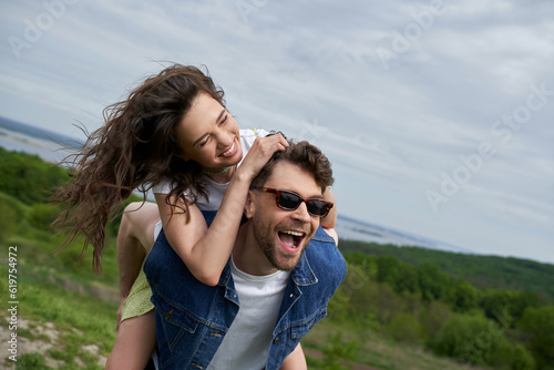 Positive brunette woman piggybacking on trendy boyfriend in sunglasses and denim vest while having fun and standing with blurred landscape at background, countryside adventure and love story