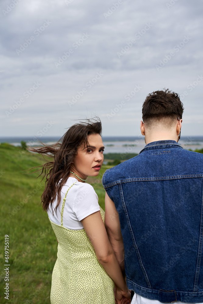 Fashionable brunette woman in summer sundress looking at camera and holding hand of boyfriend in denim vest and standing on blurred green field, couple in love enjoying nature, tranquility Fashionable brunette woman in summer sundress looking at camera and holding hand of boyfriend in denim vest and standing on blurred green field, couple in love enjoying nature, tranquility