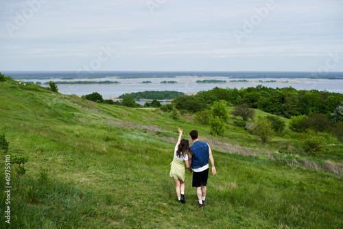 Back view of stylish couple in summer outfits holding hands and standing on grassy field with scenic view and cloudy sky at background, couple in love enjoying nature, tranquility