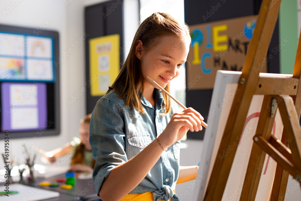 Fototapeta premium Happy caucasian schoolgirl painting using brush and easel in school art class