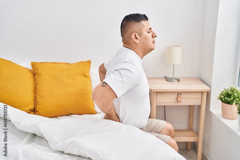Young latin man suffering for back injury sitting on bed at bedroom Young latin man suffering for back injury sitting on bed at bedroom