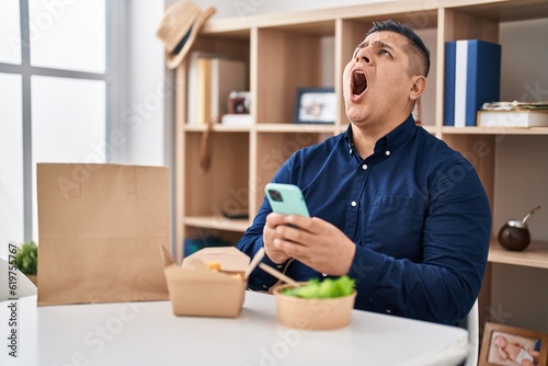 Hispanic young man eating take away food using smartphone angry and mad screaming frustrated and furious, shouting with anger looking up.