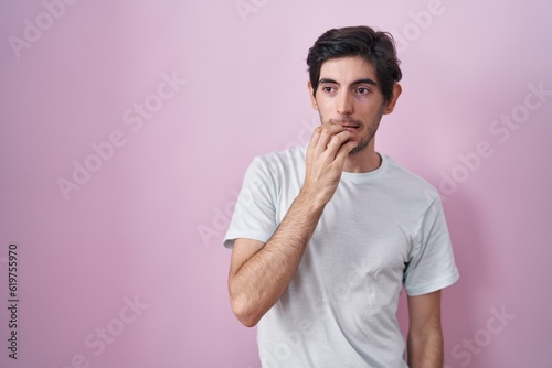 Young hispanic man standing over pink background looking stressed and nervous with hands on mouth biting nails. anxiety problem.