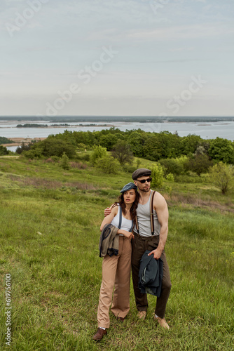 Fashionable man in sunglasses and newsboy cap hugging brunette girlfriend and holding jacket while spending time and standing on grassy field  trendy couple in the rustic outdoors