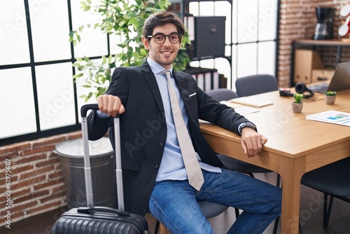 Young hispanic man business worker waiting for travel holding suitcase at office