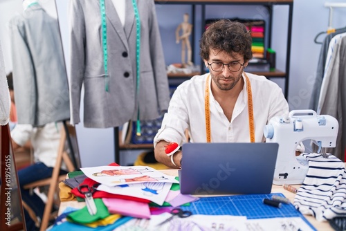 Young hispanic man tailor using laptop at clothing factory