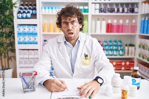Hispanic young man working at pharmacy drugstore in shock face, looking skeptical and sarcastic, surprised with open mouth
