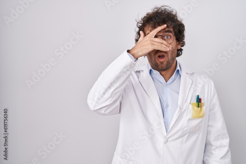 Hispanic young man wearing doctor uniform peeking in shock covering face and eyes with hand, looking through fingers with embarrassed expression.