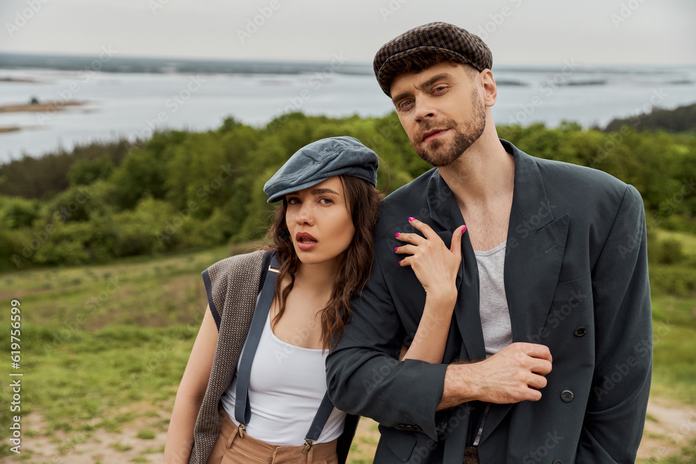 Fashionable brunette woman in suspenders and newsboy cap hugging bearded boyfriend in jacket and looking at camera together with blurred landscape at background, fashionable couple in countryside