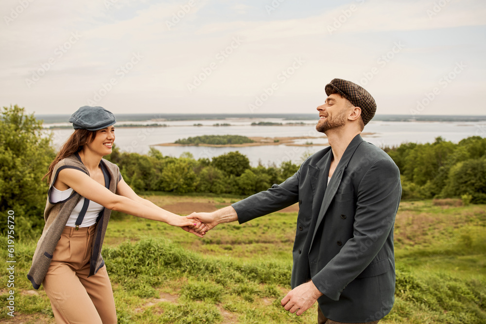 Positive and fashionable brunette woman in newsboy cap and vest holding hand of bearded boyfriend in jacket while standing with scenic landscape at background, fashionable couple in countryside Positive and fashionable brunette woman in newsboy cap and vest holding hand of bearded boyfriend in jacket while standing with scenic landscape at background, fashionable couple in countryside