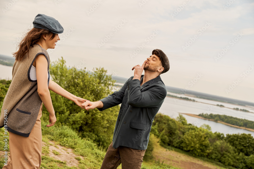 Fashionable man in newsboy cap and jacket showing secret gesture and holding hand of cheerful girlfriend while standing with nature at background, fashion-forwards in countryside Fashionable man in newsboy cap and jacket showing secret gesture and holding hand of cheerful girlfriend while standing with nature at background, fashion-forwards in countryside