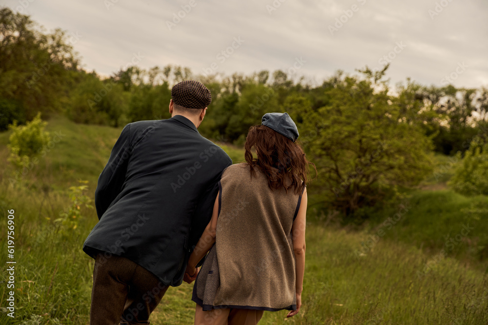 Back view of fashionable couple in newsboy caps and vintage outfits holding hands and walking together on blurred field with landscape at background, stylish couple in rural setting