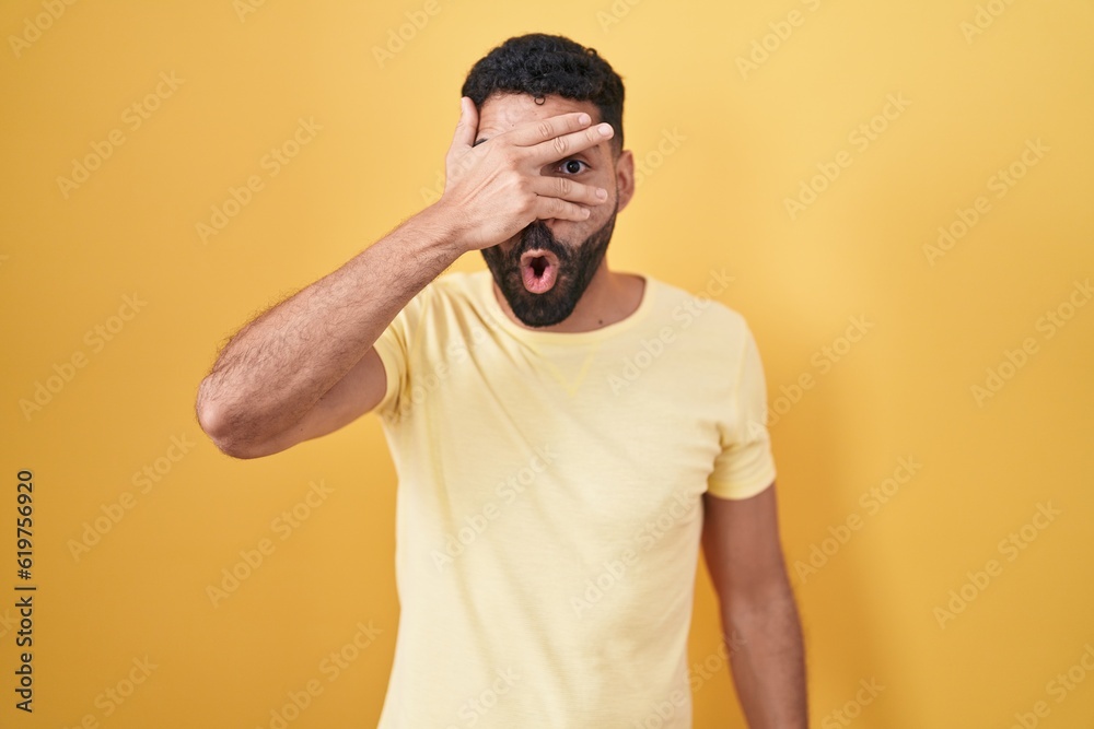 Hispanic man with beard standing over yellow background peeking in shock covering face and eyes with hand, looking through fingers with embarrassed expression.