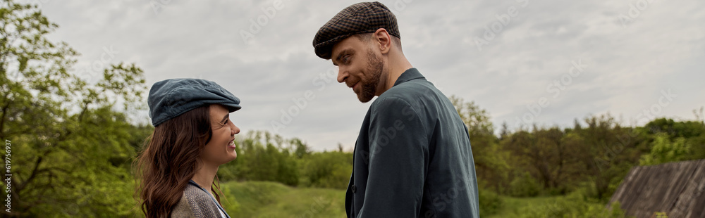 Cheerful brunette and stylish woman in newsboy cap looking at bearded boyfriend in jacket while standing together with landscape and overcast at background, stylish couple in rural setting, banner Cheerful brunette and stylish woman in newsboy cap looking at bearded boyfriend in jacket while standing together with landscape and overcast at background, stylish couple in rural setting, banner