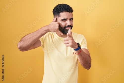 Hispanic man with beard standing over yellow background smiling doing talking on the telephone gesture and pointing to you. call me. © Krakenimages.com