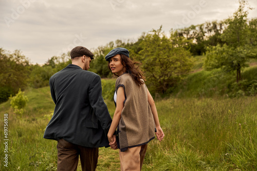 Fashionable brunette woman in vest and newsboy cap looking at camera and holding hand of bearded boyfriend and walking with landscape at background  stylish couple in rural setting