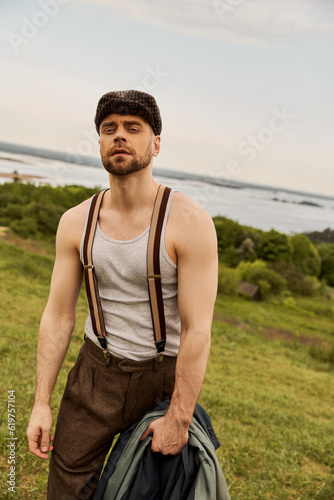 Portrait of fashionable and bearded man in suspenders and newsboy cap holding jacket and looking at camera while standing with blurred landscape at background, fashion-forward in countryside