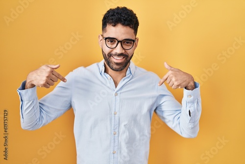 Hispanic man with beard standing over yellow background looking confident with smile on face, pointing oneself with fingers proud and happy.