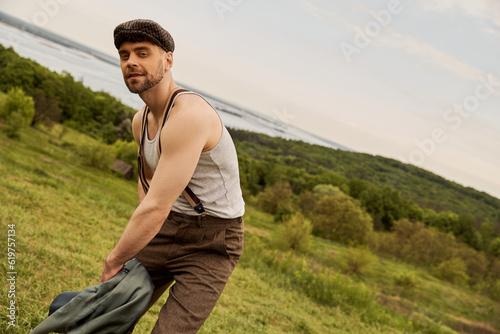 Trendy bearded man in newsboy cap and suspenders holding jacket and looking at camera while posing in vintage outfit with landscape at background, scenic countryside concept