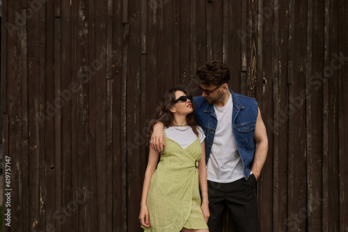 Fashionable brunette man in sunglasses and denim vest hugging smiling girlfriend in sundress and holding hand in pocket while standing near wooden house, countryside exploration concept
