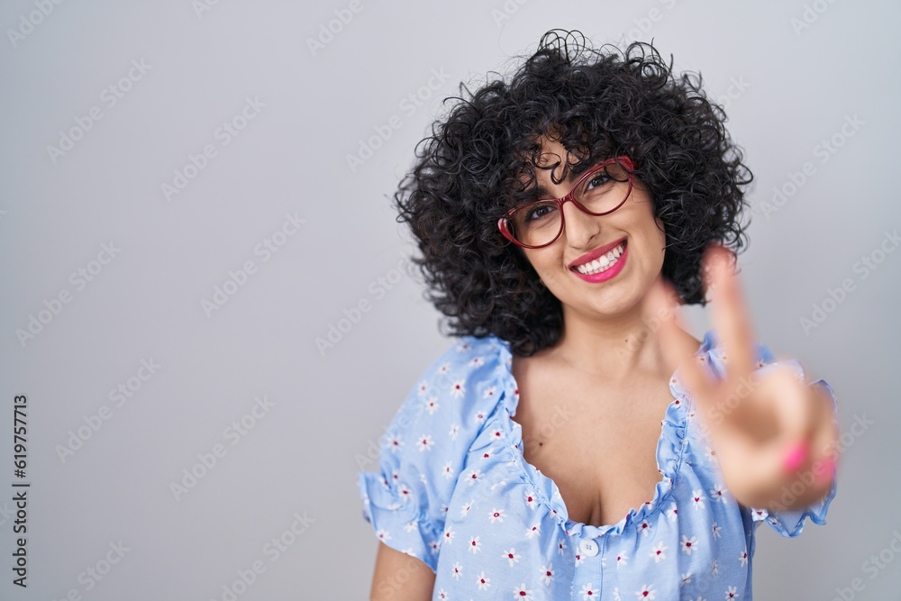 Young brunette woman with curly hair wearing glasses over isolated background smiling looking to the camera showing fingers doing victory sign. number two. Young brunette woman with curly hair wearing glasses over isolated background smiling looking to the camera showing fingers doing victory sign. number two.