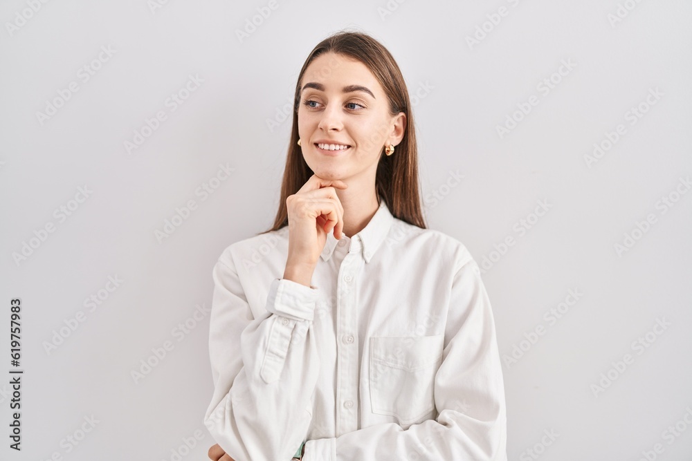 Young caucasian woman standing over isolated background with hand on chin thinking about question, pensive expression. smiling and thoughtful face. doubt concept. Young caucasian woman standing over isolated background with hand on chin thinking about question, pensive expression. smiling and thoughtful face. doubt concept.