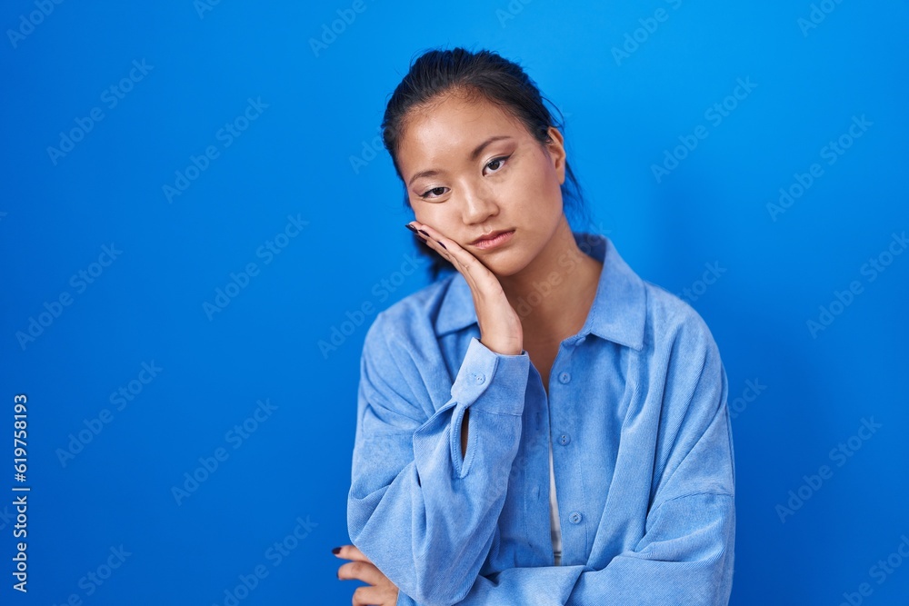 Asian young woman standing over blue background thinking looking tired and bored with depression problems with crossed arms.