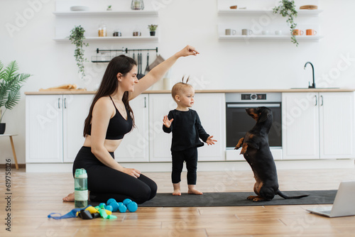 Foto Smiling female in activewear holding dog's treat in air while cute black Dachshund sitting on hind legs near baby girl