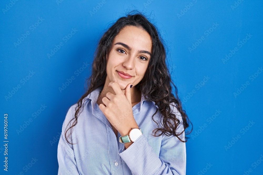 Young brunette woman standing over blue background looking confident at the camera smiling with crossed arms and hand raised on chin. thinking positive. Young brunette woman standing over blue background looking confident at the camera smiling with crossed arms and hand raised on chin. thinking positive.