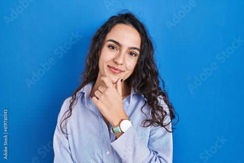 Young brunette woman standing over blue background looking confident at the camera smiling with crossed arms and hand raised on chin. thinking positive.