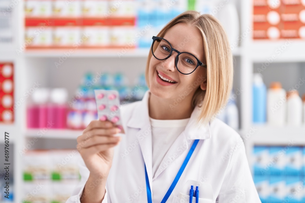 Young blonde woman pharmacist smiling confident holding pills tablet at pharmacy